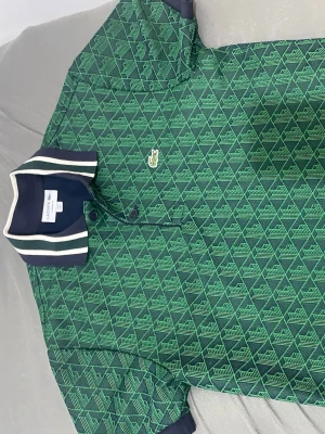 Grön mönstrad piké från Lacoste - Säljer en grön pikétröja från Lacoste med geometriskt mönster och klassisk krage i mörkblått med vita ränder. Tröjan har korta ärmar och den ikoniska krokodilloggan på bröstet. Perfekt för dig som vill ha en stilren och sportig look.