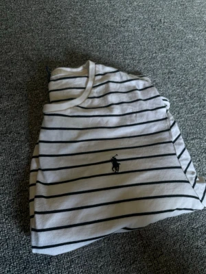 Randig t-shirt från Polo Ralph Lauren - Säljer en vit t-shirt från Polo Ralph Lauren med svarta horisontella ränder och den klassiska loggan broderad på bröstet. Modellen är custom slim fit och tillverkad i mjuk bomull. Perfekt för en clean och stilren look.
