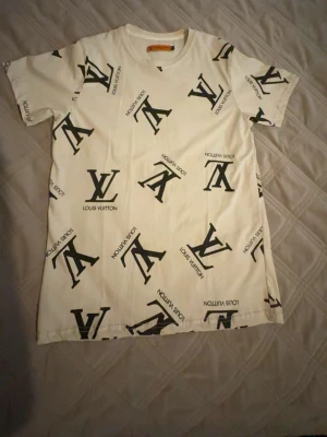 Vit Louis Vuitton t-shirt med logga - Säljer en vit t-shirt från Louis Vuitton med svart LV-logga tryckt över hela tröjan. T-shirten har rund halsringning och korta ärmar. Materialet känns mjukt och skönt, perfekt för en clean och trendig look.