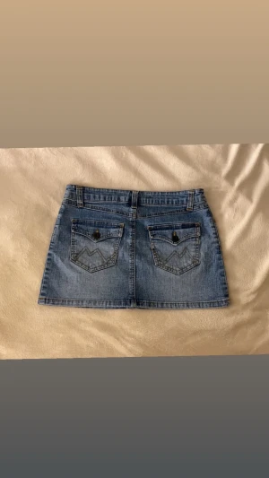 Blå jeanskjol med fickor - Säljer en klassisk blå jeanskjol i kort modell med fem fickor och bälteshällor. Kjolen har två bakfickor med dekorativa sömmar och knappdetaljer. Perfekt till vardags för en avslappnad och trendig look.