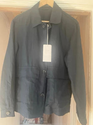Marinblå overshirt med knappar SMALL storlek - Stilren marinblå overshirt med klassisk krage och knäppning framtill. Jackan har raka linjer, stora fickor och är tillverkad i ett lätt material som passar perfekt för lager-på-lager. Långärmad modell med enkel design som funkar till många olika stilar. Aldrig använd. Prislappen sitter kvar. Felköp. Storlek small  
