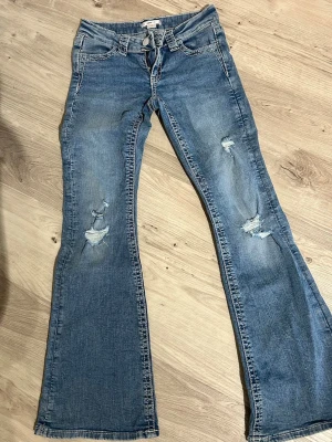 Blå bootcut jeans med slitningar - Säljer ett par blå bootcut jeans med slitningar på framsidan och broderade bakfickor. Jeansen har låg midja, klassisk femficksmodell och är gjorda i denim. Perfekta för en avslappnad och trendig look.