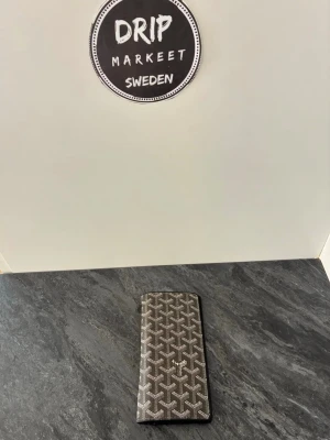 Svart Goyaard plånbok med mönster - Snygg svart plånbok från Goyaard med det klassiska vita och gråa grafiska mönstret. Slimmad och avlång modell som rymmer kort och sedlar. Märkets logga syns framtill. Perfekt accessoar för dig som gillar exklusiv design.