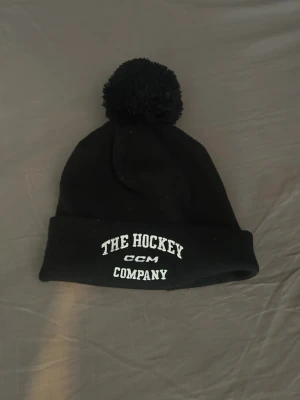 Svart CCM mössa - Säljer en svart mössa från CCM med vit brodyrtext 'The Hockey Company' framtill och en stor boll på toppen. Perfekt för kalla dagar och hockeyfans som vill hålla stilen. Klassisk design med uppvikt kant.