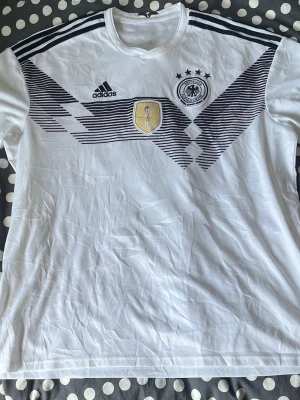 Tysklands matchtröja Adidas 2XL - Vit Tyskland matchtröja från Adidas med svarta detaljer och mönster över bröstet, fyra stjärnor och VM-märke. Klassisk rund hals och korta ärmar. Perfekt för fotbollsfans som vill visa sitt stöd för landslaget.