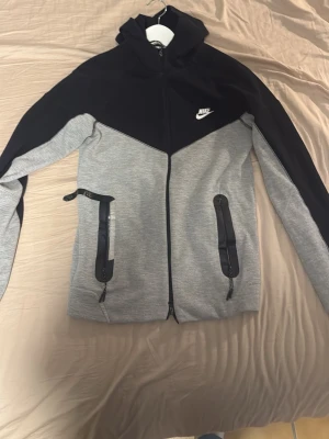 Nike grå och svart hoodie - Nike hoodie i grått och svart med dragkedja framtill och två fickor med svarta detaljer. Klassisk huva och Nike-logga på bröstet. Tillverkad i mjukt material som passar perfekt till en avslappnad stil.