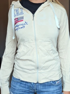Lexington hoodie - Säljer en beige hoodie med dragkedja och huva. Framtill finns tryck med texten lex och en amerikansk flagga samt blå brodyr. Hoodien har fickor på sidorna och vita snören i huvan. Perfekt till jeans för en avslappnad look.