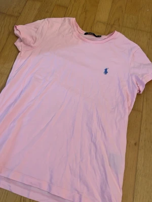Ljusrosa t-shirt från Polo Ralph Lauren - Säljer en ljusrosa t-shirt från Polo Ralph Lauren med klassisk blå logga broderad på bröstet. T-shirten har korta ärmar, rund halsringning och är gjord i mjuk bomull. Perfekt för en avslappnad och stilren look.