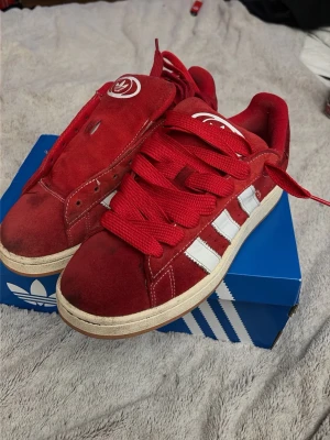 Röda Adidas Campus sneakers - Säljer ett par röda Adidas Campus sneakers med klassiska vita ränder på sidorna och tjocka röda skosnören. Skorna har låg profil och ovandel i mocka, samt Adidas-logga på plösen. Snygg retrostil som passar till många outfits. Ett skosnöre till skorna saknas men skickas med två röda extra snören. Låda skickas med.