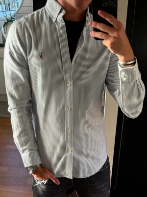 Randig skjorta från Polo Ralph Lauren - Säljer en klassisk blåvit randig skjorta från Polo Ralph Lauren i slim fit. Skjortan har långa ärmar, knappar framtill och button-down krage. Tillverkad i bomull och har den ikoniska loggan broderad på bröstet. Perfekt för en clean och stilren look.
