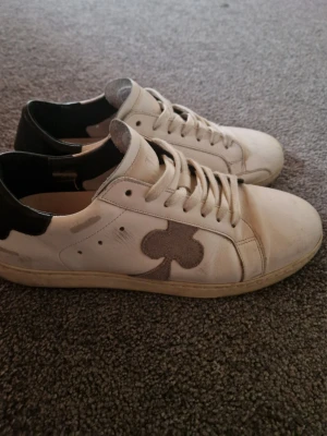 The Gallerist - Säljer ett par vita sneakers med svart häl och en grå klöverdetalj på sidan. Skorna har klassisk låg profil, vita snören och är tillverkade i läder. Perfekta för dig som gillar stilrena och unika detaljer på dina skor.