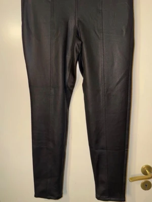 Svarta leggings i fuskskinn - Snygga svarta leggings i fuskskinn med hög midja och sömdetaljer längs benen. Tight passform som ger en cool look och passar perfekt till oversized tröjor eller crop tops. Materialet har en lätt glansig finish som ger extra edge till outfiten.