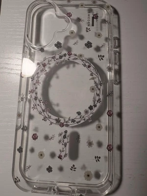 Genomskinligt mobilskal med blommor - Snyggt genomskinligt mobilskal med blommönster och MagSafe-kompatibilitet. Skalets kanter är rundade och det har ett hjärtformat kamerauttag. Passar perfekt för dig som vill ha ett unikt och stilrent skydd till din mobil.