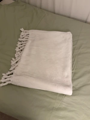 Krämvit filt från Hemtex  - Säljer en vit, supermjuk pläd med dekorativa tofsar längs ena kanten. Perfekt att ha i soffan eller på sängen för extra mysfaktor. Materialet känns fluffigt och lent mot huden. Enkel och stilren design som passar in i de flesta hem.