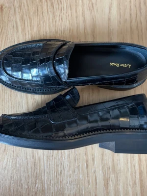 Svarta loafers i läder med reptilmönster - Snygga svarta loafers med glansigt reptilmönster i läder. Skorna har en chunky sula och klassisk pennyloafers-design. Perfekta för dig som vill ha en stilren men ändå unik look. Nypris 1100 kr.