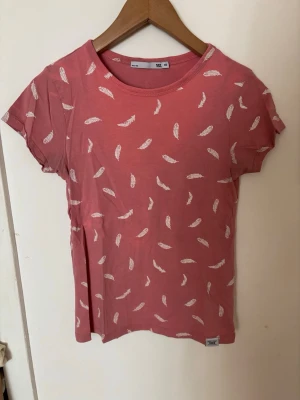 Rosa t-shirt med fjädermönster - Säljer en rosa t-shirt med vita fjädrar tryckta över hela plagget. T-shirten har rund halsringning och korta ärmar🩷