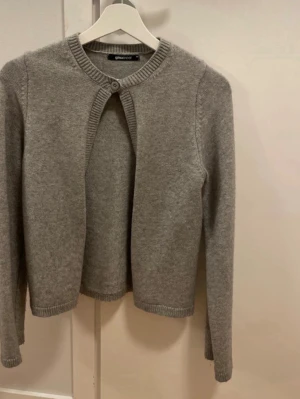 Grå kofta från Gina Tricot - Mysig grå kofta från Gina Tricot med rund halsringning och en knapp upptill. Koftan har långa ärmar och ribbade muddar vid ärmslut och nederkant. Perfekt att slänga över en topp för extra värme.