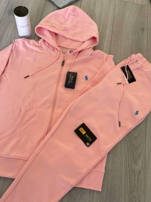 Rosa loungewear-set från Polo Ralph Lauren - Säljer ett mjukt och bekvämt loungewear-set i ljusrosa från Polo Ralph Lauren. Setet består av en hoodie med dragkedja och fickor samt matchande byxor med dragsko i midjan. Båda plaggen har den klassiska blå loggan broderad på bröstet och benet.