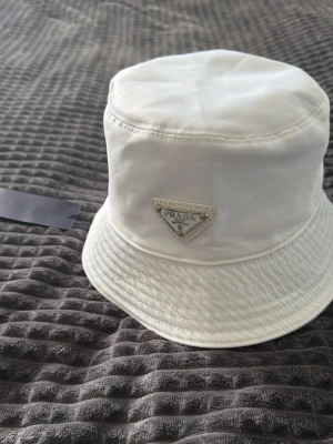 Vit bucket hat från Prada - Snygg vit bucket hat från Prada med klassisk triangulär logga framtill. Hatten är tillverkad i ett glansigt material som ger en exklusiv känsla och har en mjuk, rundad form. Perfekt accessoar för att lyfta din outfit med en lyxig touch.