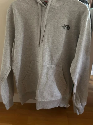 Grå hoodie från The North Face - Säljer en stilren grå hoodie från The North Face med klassisk logga på bröstet. Hoodien har känguruficka, dragsko i huvan och ribbade muddar. Perfekt för chill dagar eller när du vill ha en enkel och snygg look.