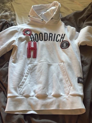 Vit hoodie från Hoodrich med patchar - Säljer en vit hoodie från Hoodrich med svart text, röda och svarta patchar samt stor ficka framtill. Hoodien har huva med dragsko och ribbade muddar. Perfekt för dig som gillar streetwear och vill sticka ut med snygga detaljer.