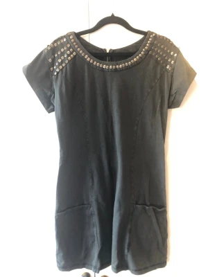 VILA Klänning Sienna m. Nitar / Rockig – Stl S - Cool klänning eller tunika i mjuk jersey från VILA. Rakt skuren modell med två funktionella fickor fram och dragkedja bak. Rockig stil med metall nitar vid axlar och hals. Klänningen är något urtvättad i tyget. Inga nitar saknas, men två nitar på vänster axel har börjat lossna och sitter löst (se bilder). Storlek: S Mått: Längd ca 79 cm, axelbredd ca 39 cm Material: 97% bomull, 3% spandex Skick: Begagnat skick (se beskrivning)