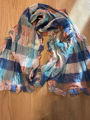 Randig pastellfärgad sjal - Säljer en tunn och luftig sjal med breda ränder i pastellfärger som blått, persika, beige och mintgrönt. Sjalen har en skrynklig struktur och vågiga kanter som ger en avslappnad vibe. Perfekt att slänga över axlarna för en färgklick till din outfit.