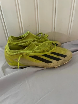 Gula Adidas fotbollsskor med snörning - Säljer ett par gula Adidas fotbollsskor med svarta ränder på sidan och snörning. Skorna har en strumpliknande öppning och är designade för bra grepp på planen. Perfekta för dig som vill sticka ut på fotbollsplanen.