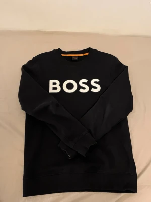 Svart sweatshirt från BOSS - Snygg svart sweatshirt från BOSS med stor vit logga framtill. Tröjan har rund halsringning, ribbade muddar vid ärmslut och nederkant samt en orange detalj på insidan av kragen. Perfekt för en stilren och avslappnad look.