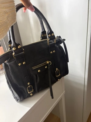 Svart handväska med gulddetaljer - Snygg svart handväska i skinnimitation med guldfärgade detaljer och dragkedjor. Väskan har dubbla handtag, dekorativa spännen och en liten ficka framtill med dragkedja. Perfekt storlek för det viktigaste och ger en cool vibe till din outfit. Andvänt få gånger, så är i mycket är skick!