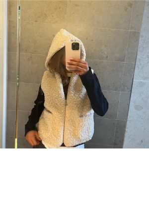 Beige teddyväst med huva - Mysig beige teddyväst med dragkedja framtill och stor huva. Västen har två rymliga fickor och är gjord i fluffigt, mjukt material som ger en avslappnad och trendig look. Perfekt att slänga över en hoodie eller tröja för extra värme.