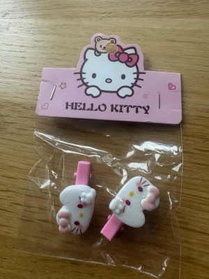 Hello Kitty hårspännen rosa och vit - Söta hårspännen med Hello Kitty-motiv i vitt och rosa. Spännena har form av Hello Kittys ansikte med rosa rosett och detaljer i gult och rött. Perfekta för att piffa upp frisyren med en gullig touch.