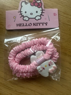 Hello Kitty rosa hårsnoddar - Två söta rosa hårsnoddar med Hello Kitty-motiv. Snoddarna är täckta av glansigt tyg och har en dekorativ Hello Kitty-detalj i plast. Perfekt för att piffa upp din frisyr med en gullig touch.