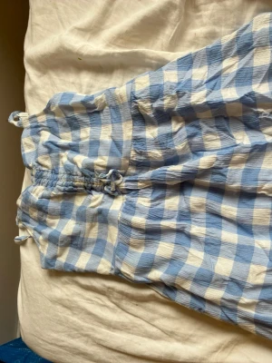 Blåvit rutig sommarklänning - Söt klänning med blåvita rutor och smock upptill. Klänningen har tunna axelband och är gjord i ett lätt, krinklat tyg som känns luftigt. Perfekt för varma dagar och ger en avslappnad, trendig look.
