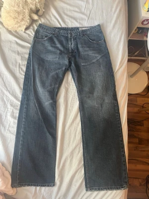 Blå raka jeans med kontrastsömmar - Säljer ett par klassiska blå jeans med rak passform och kontrastsömmar på bakfickorna. Jeansen har fem fickor och normal midja. Perfekta för dig som gillar en avslappnad och tidlös stil. Materialet är denim i bomull.