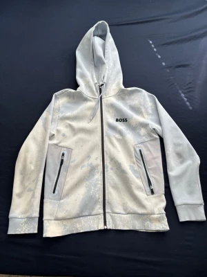 Vit/blå hoodie med dragkedja från BOSS - Snygg vit/blå fläckig hoodie från BOSS med dragkedja framtill och två fickor med svarta dragkedjor. Hoodien har ett unikt blekt mönster och justerbar huva med snören. Tillverkad i mjukt material som känns skönt mot huden. Perfekt för en avslappnad streetwear-look.