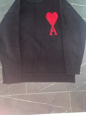 Svart stickad tröja från Ami - Snygg svart stickad tröja från Ami med röd hjärtlogga och bokstaven A på bröstet. Tröjan har ribbade muddar vid ärmslut och nederkant samt rund halsringning. Perfekt för dig som gillar stilrena plagg med en twist.