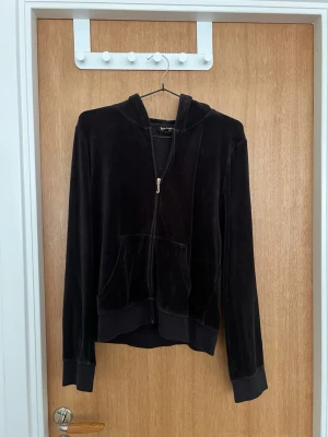 Svart velourhoodie med dragkedja - Mjuk och skön svart hoodie i velour med dragkedja framtill och känguruficka. Klassisk huva och ribbade muddar vid ärmslut och nederkant. Perfekt för en avslappnad och trendig look.