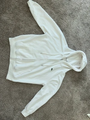 Vit zip hoodie från Lacoste - Snygg vit hoodie från Lacoste med dragkedja framtill och klassisk grön krokodillogga på bröstet. Hoodien har huva med dragsko och två fickor framtill. Tillverkad i mjukt bomullsmaterial som känns skönt mot huden.