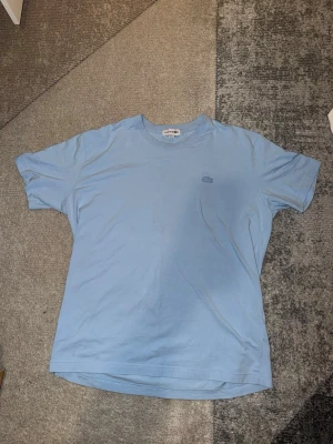 Ljusblå T-shirt - Säljer en ljusblå t-shirt med den klassiska krokodilloggan broderad på bröstet. T-shirten har rund hals och korta ärmar, tillverkad i mjuk bomull för en skön och avslappnad stil.