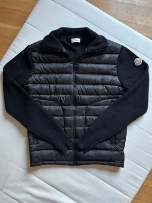Svart pufferjacka från Moncler - Snygg svart pufferjacka från Moncler med stickade ärmar och krage. Jackan har quiltad front i glansigt material, dragkedja framtill och två fickor med dragkedja. Moncler-logga på vänster ärm ger en exklusiv touch. Perfekt för kalla vinterdagar.