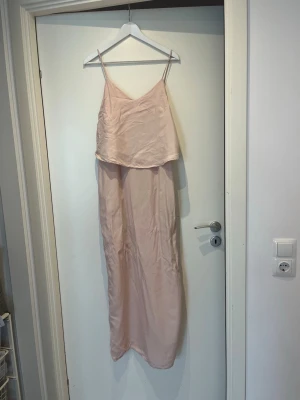 Ljusrosa långklänning Stylein - En ljusrosa lång klänning från Stylein i storlek M.  Klänningen har slits på baksidan (obs, lite av sömmen har gått upp precis där, syns på bild 3)  Klänningen har fickor på båda sidor! Dragkedja sitter på ena sidan.  Smala axelband som är justerbara.  Mätte klänningen och total längd fast utan axelbanden inräknade är 130 cm. Material: yttertyg 55% cupro* (*fibrer från bomullsfröerna som vanligtvis annars kasseras när man tillverkar bomullstyg), 45% viskos. Innertyg är 100% polyester 