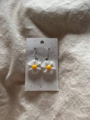 Örhängen med prästkrageblommor - Säljer ett par söta örhängen med vita prästkrageblommor och gul mitt. Hängena är i metall och blommorna har en tydlig 3D-effekt som gör dem extra charmiga. Perfekta för dig som gillar blommiga accessoarer och vill sticka ut lite extra.