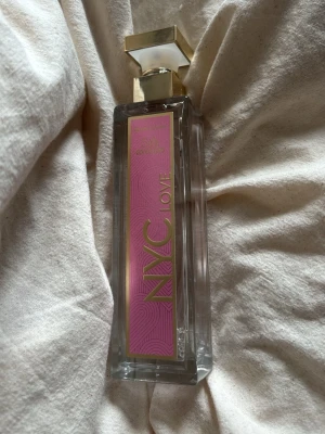 NYC Love parfym med guldlock - NYC Love parfym i en hög, rektangulär glasflaska med guldigt lock och detaljer. Framsidan har en rosa etikett med guldtext. Flaskan är genomskinlig och elegant, perfekt för dig som gillar snygg design på parfymflaskor.