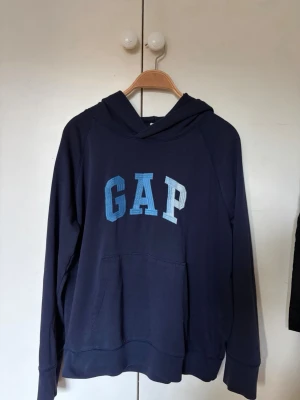 Mörkblå hoodie GAP - Mörkblå hoodie från GAP med stor logga i blå toner på bröstet. Klassisk känguruficka framtill och huva. Tillverkad i mjukt bomullsmaterial, perfekt för en avslappnad och cool stil. Axelbredd är 43cm, längd är 64cm