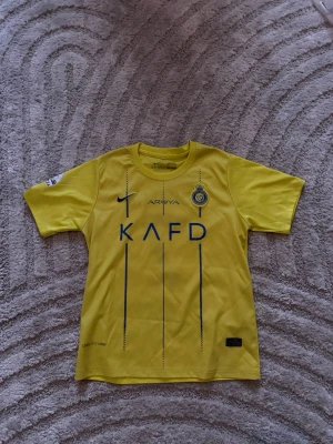 Gul Al Nassr Ronaldo matchtröja Nike - Snygg gul Al Nassr matchtröja från Nike med Ronaldo 7 på ryggen. Tröjan har korta ärmar, klubbmärke och sponsorloggor framtill samt tryck på ärmen. Tillverkad i lätt och ventilerande material, perfekt för fotboll eller träning.