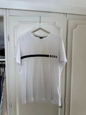 Vit BOSS t-shirt med svart tryck - Snygg vit t-shirt från BOSS med svart vågig linje och logga på bröstet. Klassisk rund hals och korta ärmar. Tillverkad i mjuk bomull för skön känsla och enkel stil.
