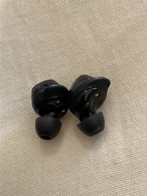 Svarta trådlösa in-ear hörlurar Samsung  - Snygga svarta trådlösa in-ear hörlurar med rundad form och mjuka silikonpluggar. Perfekta för musik, poddar eller samtal på språng. Smidig och kompakt design som passar i fickan. Märke och modell syns inte tydligt på bilden.