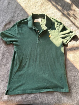 Grön pikétröja från Lyle & Scott - Grön pikétröja från Lyle & Scott med klassisk krage, två knappar och gul broderad logga på bröstet. Tillverkad i mjuk bomull och har en clean, avslappnad passform. Nypris 499 Kr