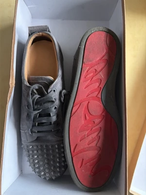 Gråa Louboutin sneakers med nitar - Säljer ett par gråa sneakers från Christian Louboutin i mocka med ikoniska röda sulor och coola nitar över tån. Skorna har snörning och låg profil som ger en edgy vibe. Perfekta för dig som vill sticka ut med din stil.